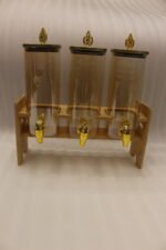 Triple 2.2L amber glass beverage dispenser set with ornate gold spigots and lids on a natural wooden stand on a marble countertop.