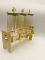 Triple 2.2L amber glass beverage dispenser set with ornate gold spigots and lids on a natural wooden stand on a marble countertop.