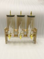 A modern kitchen counter with a three-vessel amber glass beverage dispenser set, held in a bamboo wooden stand. Each 1.8L cylinder has a gold pineapple lid and gold spigot, filled with amber liquid.