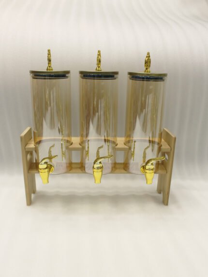 A modern kitchen counter with a three-vessel amber glass beverage dispenser set, held in a bamboo wooden stand. Each 1.8L cylinder has a gold pineapple lid and gold spigot, filled with amber liquid.