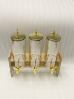 A modern kitchen counter with a three-vessel amber glass beverage dispenser set, held in a bamboo wooden stand. Each 1.8L cylinder has a gold pineapple lid and gold spigot, filled with amber liquid.