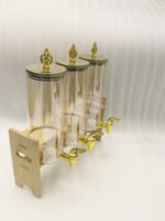 A modern kitchen counter with a three-vessel amber glass beverage dispenser set, held in a bamboo wooden stand. Each 1.8L cylinder has a gold pineapple lid and gold spigot, filled with amber liquid.