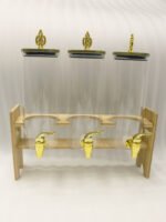 An elegant 3-tier beverage dispenser set featuring clear glass canisters with gold spigots and lids, resting on a natural bamboo stand on a white marble countertop.