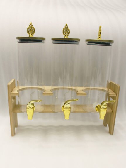 An elegant 3-tier beverage dispenser set featuring clear glass canisters with gold spigots and lids, resting on a natural bamboo stand on a white marble countertop.
