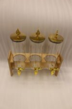 An elegant 3-tier beverage dispenser set featuring clear glass canisters with gold spigots and lids, resting on a natural bamboo stand on a white marble countertop.