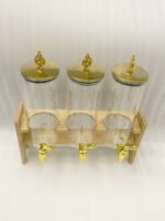 An elegant 3-tier beverage dispenser set featuring clear glass canisters with gold spigots and lids, resting on a natural bamboo stand on a white marble countertop.