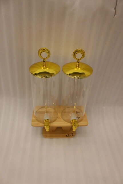 Two tall clear glass drink dispensers with gold spigots and circular gold finial lids resting on a wooden W-shaped stand.