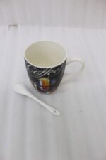 Black ceramic mug printed with vintage Coffee artwork resting on a granite kitchen counter alongside a white ceramic spoon.