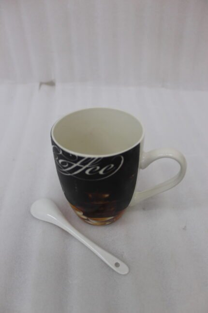 Black ceramic mug printed with vintage coffee grinder artwork next to a white ceramic spoon on a kitchen countertop.