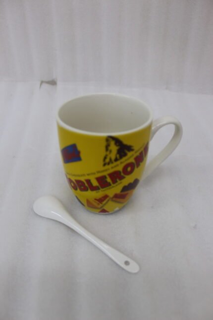 Yellow ceramic coffee mug with vintage Toblerone chocolate wrapper print and a small matching white spoon on a kitchen counter.