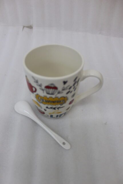 White ceramic coffee mug with colorful coffee and dessert doodle illustrations paired with a small ceramic spoon on a kitchen counter.
