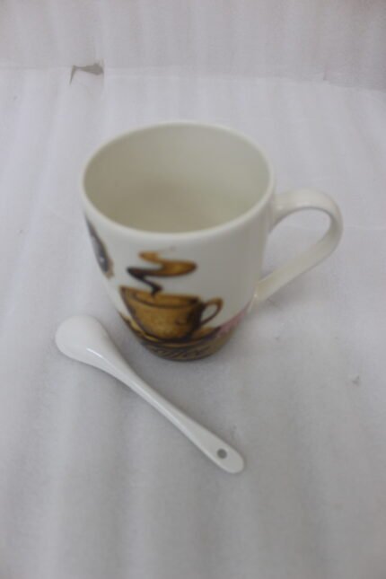 White ceramic coffee mug with vintage coffee cup graphic next to a matching white spoon on a countertop.
