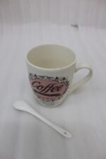 Vintage off-white ceramic coffee mug with "Coffee and then the world" text design resting on a grey countertop next to a white ceramic spoon.