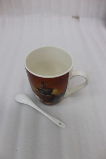 White ceramic coffee mug printed with a vintage manual coffee grinder, espresso cup, and coffee beans, sitting next to a matching white spoon on a kitchen counter.
