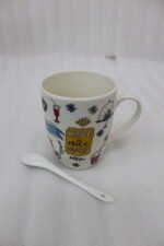White ceramic coffee mug printed with 'Have a Nice Day' doodle art next to a matching ceramic spoon on a kitchen counter.