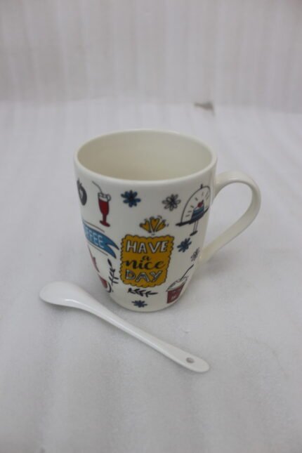 White ceramic coffee mug printed with 'Have a Nice Day' doodle art next to a matching ceramic spoon on a kitchen counter.