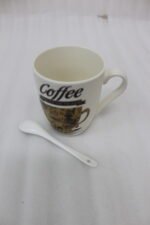 White ceramic coffee mug printed with 'Coffee and then the world' text and vintage coffee art next to a white spoon on a kitchen counter.