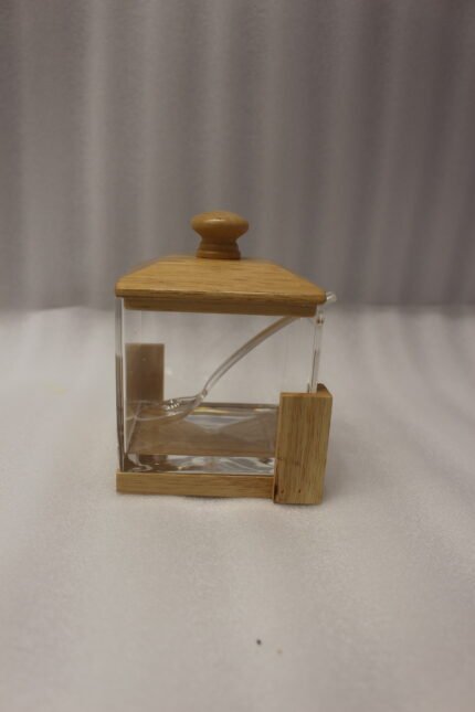 Square clear acrylic sugar pot with a natural wooden lid, clear serving spoon, and wooden base stand on a kitchen countertop.