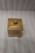 Square clear acrylic sugar pot with a natural wooden lid, clear serving spoon, and wooden base stand on a kitchen countertop.