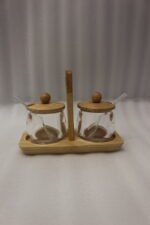 Two clear acrylic condiment jars with wooden lids and glass spoons set on a wooden stand tray with handle on a white marble countertop.
