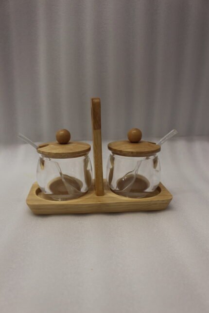 Two clear acrylic condiment jars with wooden lids and glass spoons set on a wooden stand tray with handle on a white marble countertop.