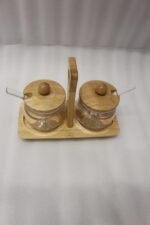 Two clear acrylic condiment jars with wooden lids and glass spoons set on a wooden stand tray with handle on a white marble countertop.