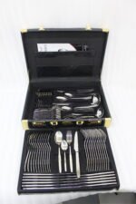 72-piece polished stainless steel cutlery set arranged on a cloth napkin featuring place settings, ladles, cake server, and tongs.