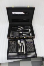 A Diverse Arrangement of Polished Silver-Toned Cutlery on a Textured Linen Mat, Featuring 12-Person Service and Loose Ladles, a Cake Server, and Tongs in the Background.