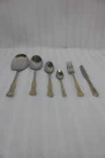 Six-piece gold accented stainless steel flatware set displayed on a linen mat including rice spoons, fork, knife, and serving spoons.
