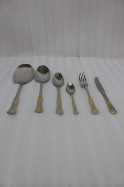 Six-piece gold accented stainless steel flatware set displayed on a linen mat including rice spoons, fork, knife, and serving spoons.