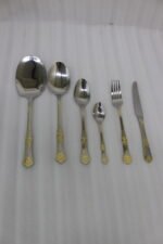 Six-piece gold accented stainless steel flatware set displayed on a linen mat including rice spoons, fork, knife, and serving spoons.