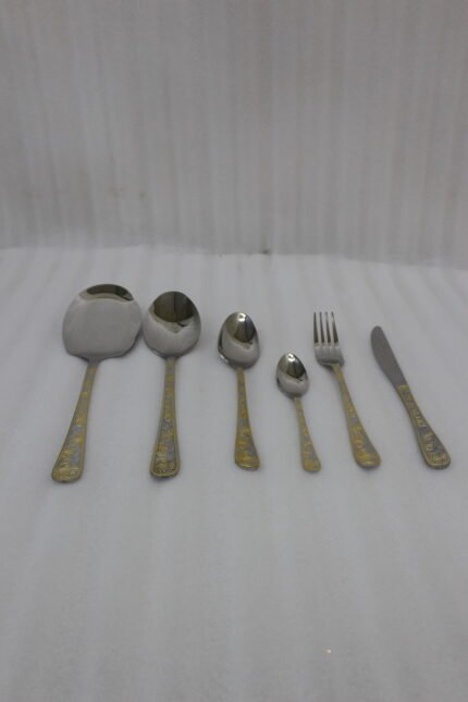Gold-embossed stainless steel cutlery set including spoons, fork, and knife arranged on a beige cloth table setting.