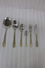 Gold-embossed stainless steel cutlery set including spoons, fork, and knife arranged on a beige cloth table setting.