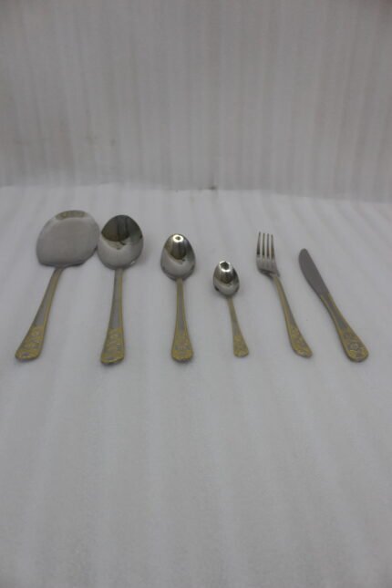 Stainless steel cutlery set with gold patterned handles featuring serving spoons, dinner forks, knives, and dessert spoons on a linen cloth.