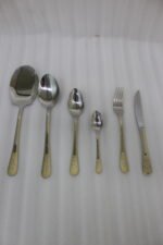 Stainless steel cutlery set with gold patterned handles featuring serving spoons, dinner forks, knives, and dessert spoons on a linen cloth.