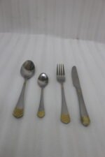 24-piece polished stainless steel cutlery set with gold filigree handle accents arranged on a beige cloth.