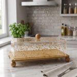Rectangular wooden cake display tray with a gold flake patterned acrylic cover and wooden ball feet on a marble countertop