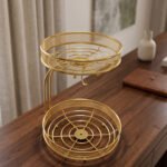Two-tier round gold wire fruit basket holder with mesh borders and integrated banana hanger hook sitting on a wooden tabletop.