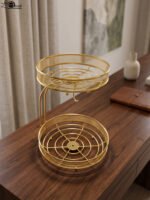 Two-tier round gold wire fruit basket holder with mesh borders and integrated banana hanger hook sitting on a wooden tabletop.