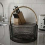 Black double-wall wire mesh fruit basket with a curved wooden handle sitting on a gray kitchen countertop.