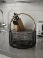 Black double-wall wire mesh fruit basket with a curved wooden handle sitting on a gray kitchen countertop.