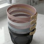 Stacked round metal wire mesh storage baskets with wooden handles in dusky pink, white, grey, and black on a marble countertop.