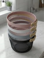 Stacked round metal wire mesh storage baskets with wooden handles in dusky pink, white, grey, and black on a marble countertop.