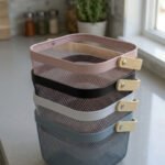 Stack of modern mesh wire storage baskets in pink, black, white, and blue with light wooden handles on a kitchen countertop.