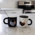 Pair of black and white ceramic couple mugs with heart accents, matching lids, and silver spoons sitting on a kitchen countertop.
