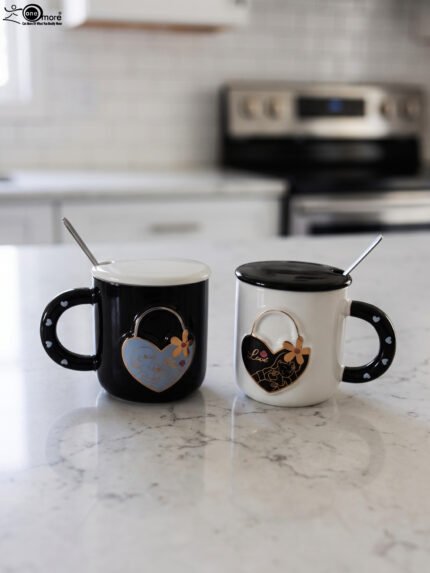 Pair of black and white ceramic couple mugs with heart accents, matching lids, and silver spoons sitting on a kitchen countertop.