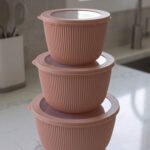Stack of three dusty pink ribbed plastic food storage bowls with clear lids on a white kitchen countertop.