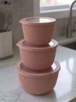 Stack of three dusty pink ribbed plastic food storage bowls with clear lids on a white kitchen countertop.
