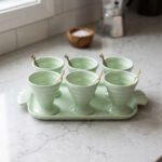 Six pastel green ceramic ice cream cups with gold leaf spoons arranged on a matching green serving tray on a kitchen countertop.