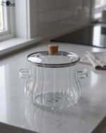 Clear ribbed glass cooking pot with wooden knob lid sitting on a white kitchen countertop